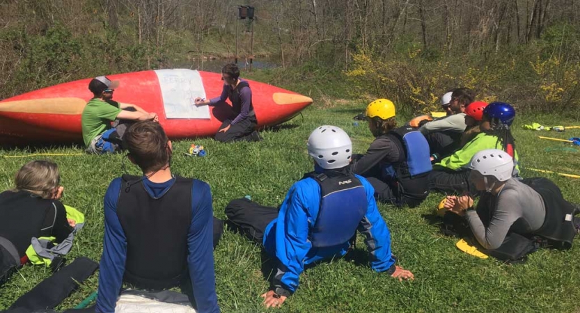 On the bottom of a tipped over canoe, an instructor points to a white sheet of paper laid across it. Students sit in a semi-circle around that canoe, listening.